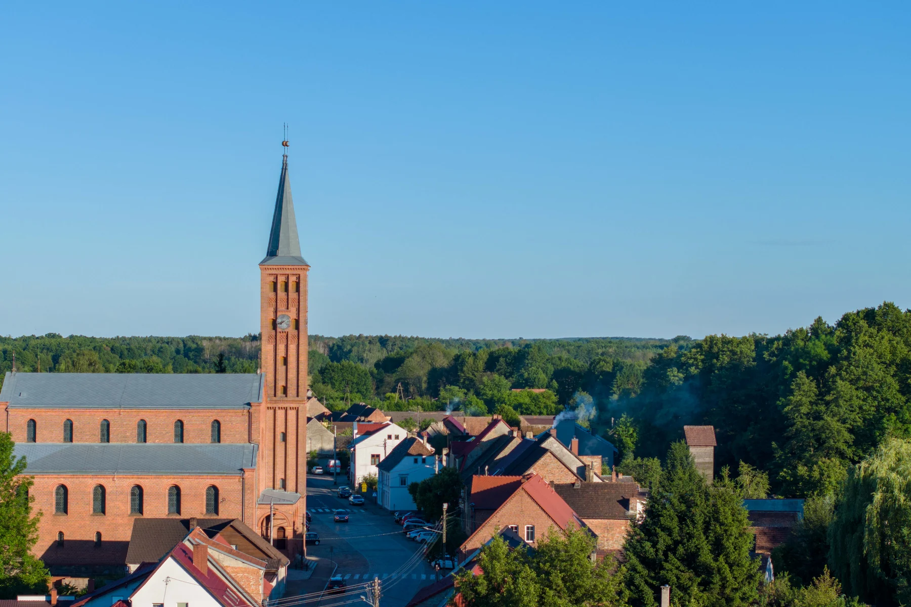 Ceglany kościół z wysoką wieżą zwieńczoną hełmem i zegarem, wokół plac i zabudowa wsi