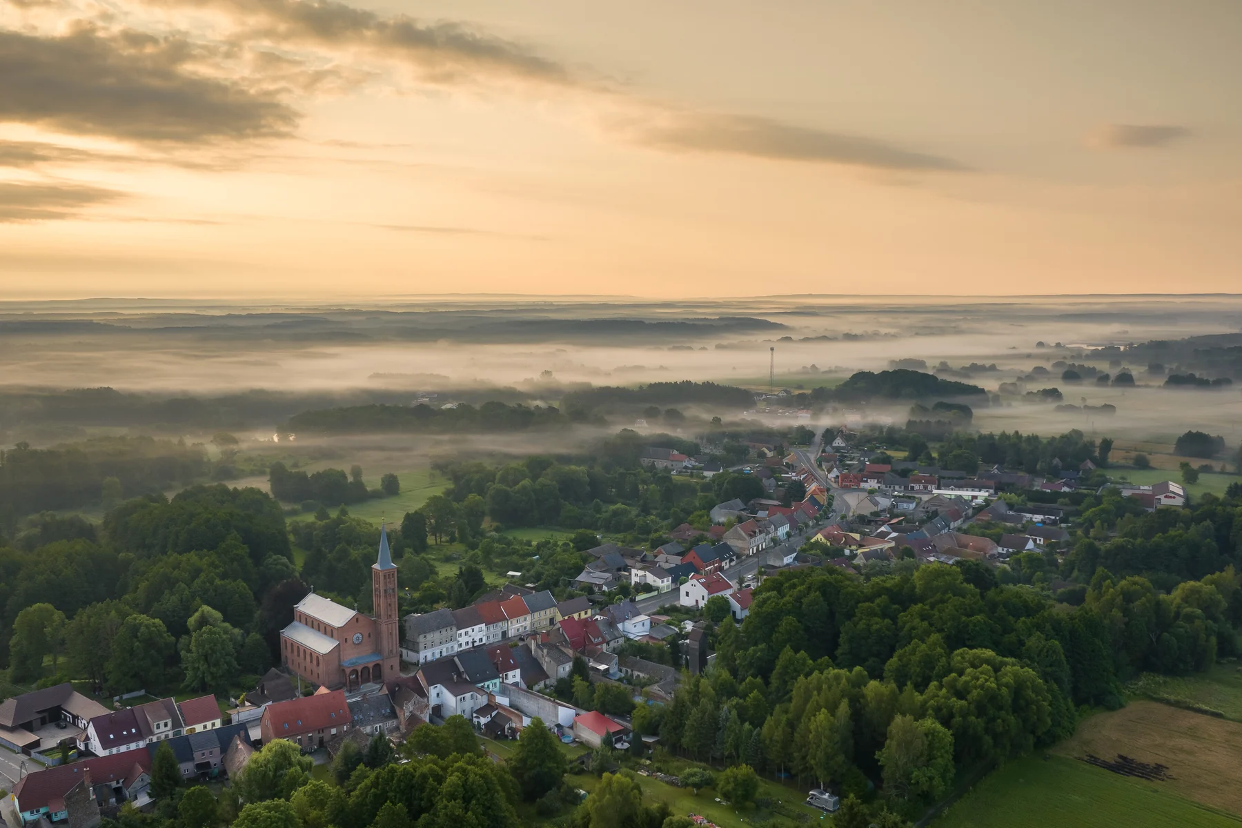 Zwarta zabudowa wsi w dolinie o świcie, ceglany kościół nad dachami, pola i pasma lasu we mgle