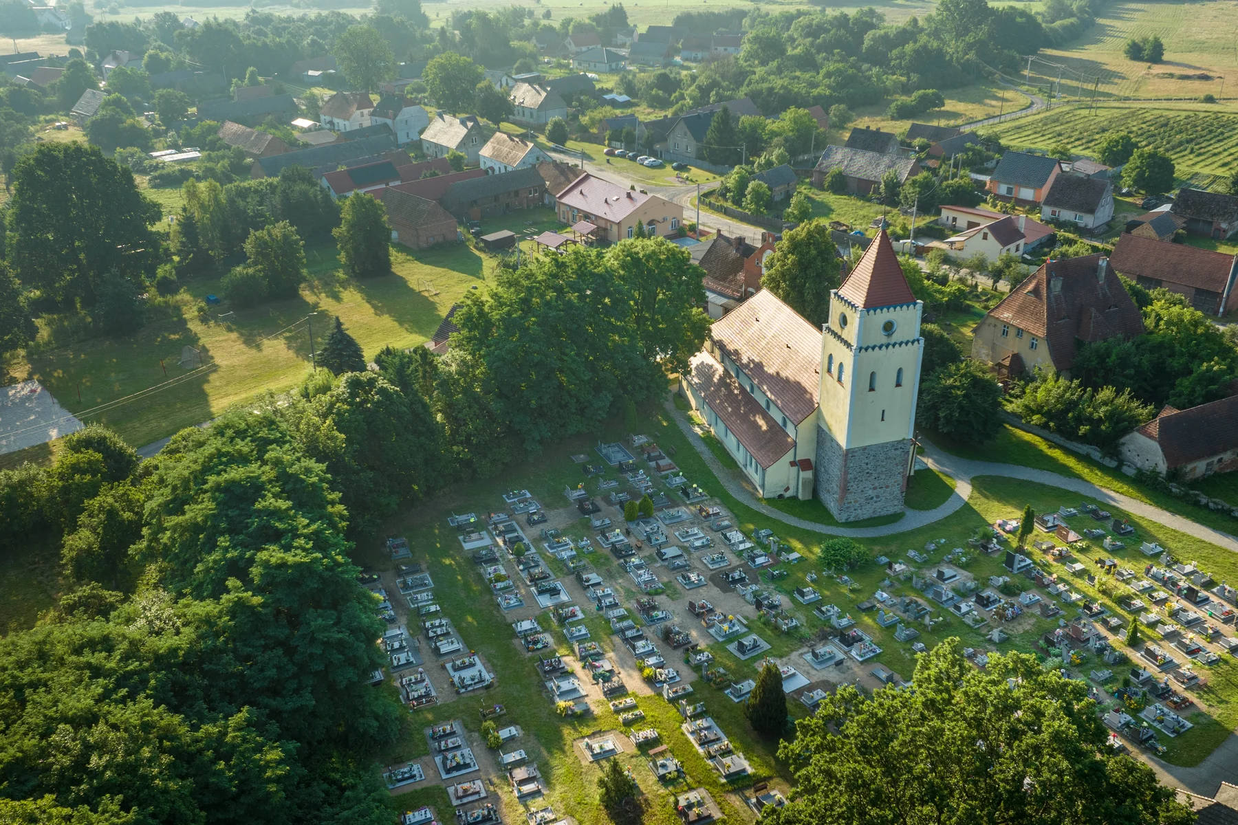 Kościół z kamienną dolną częścią wieży i otynkowaną górną, groby wokół świątyni na tej samej działce, w tle zabudowa wsi i pola
