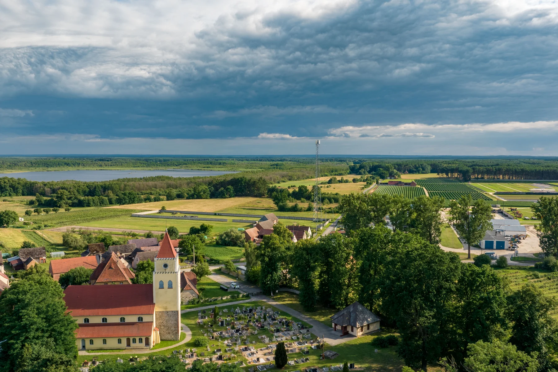 Otynkowany kościół z wieżą zwieńczoną czerwonym hełmem, obok wydzielona kwatera cmentarna za szpalerem drzew, dalej pola i tafla wody na horyzoncie